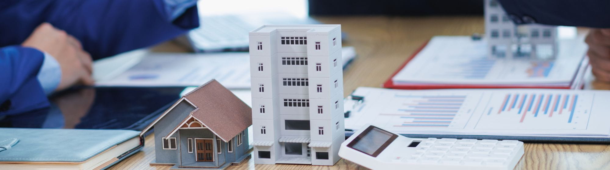 Models of a house and an apartment building placed on a table with financial documents, a calculator, and charts during a real estate investment meeting.