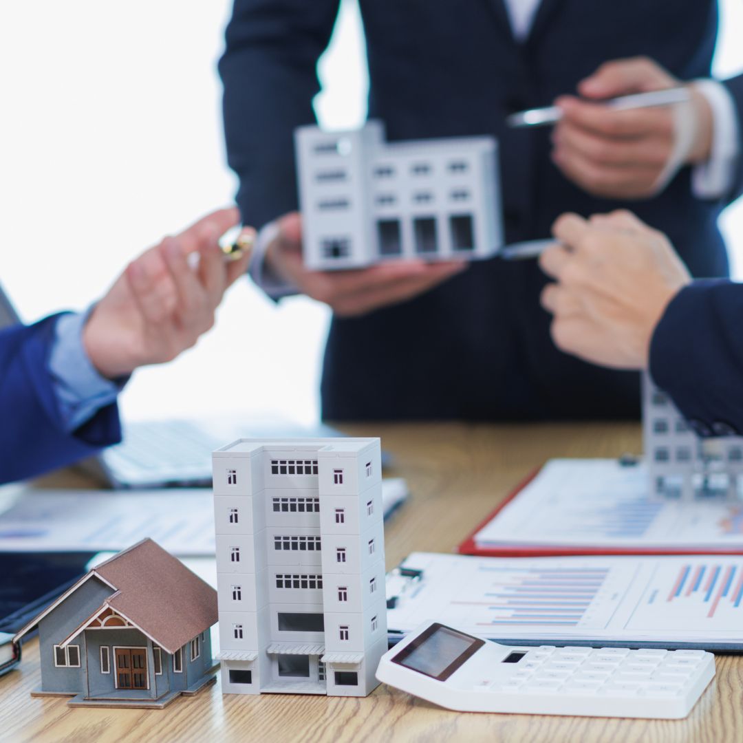 Réunion d'investissement immobilier avec des maquettes de bâtiments, une calculatrice et des documents financiers sur une table, tandis que des hommes en costume discutent en arrière-plan.